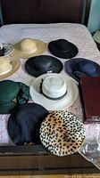 Photo showing multiple ladies hats on a bed: including five summer weight hats, five dressy hats and two berets of various colors and materials, also a dark wooden jewelry box.