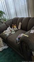 Front and angled side view of worn upholstered love seat with soft textured fabric showing some wear. Decorative figurines and miscellaneous items present but not included.