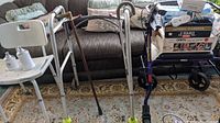 Photo showing a white plastic shower chair with backrest, two wooden canes with curved handles, a silver metal walker without wheels, and a blue walker with wheels.