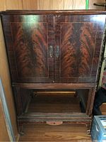 Front view of vintage radio cabinet showing two doors with metal handles, wood grain finish, and bottom compartment open without shelf.