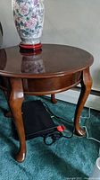 View of one round wooden side table with glossy top, drawer, and cabriole legs in Queen Anne style on green carpet with a floral vase on top.