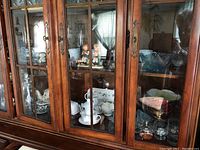 Wooden glass-front cabinet with multiple items including tea cups, Wedgwood china, wine glasses, serving dishes, and decorative figurines.