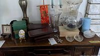 View of various glass and decorative items on a dark wooden dresser including pressed glass heart dish, engraved egg trinket box, two vases, two candle holders, empty boxes, and other small items