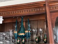 Front view of wooden china cabinet shelf holding Venetian green glass pitcher with gold trim and matching green stemmed wine glasses featuring pink rose painted bases. Surrounding glassware includes clear crystal wine and cordial glasses with cut patterns.