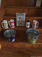 Full view of all items including four thermal beer mugs, two metal ice buckets, and a metal beer sign on wooden stairs.