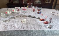 View of about 10 assorted branded beer glasses lined on a towel, showing a variety of tall glasses and goblets with logos including Miller High Life and Smirnoff.