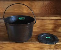 Front-side view of metal cauldron with attached handle and a separate round plate beside it, placed on wooden surface.