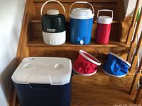 Seven beverage coolers and jugs placed on wooden stairs showing a mix of hard plastic and collapsible fabric coolers