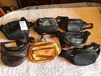 Photo of seven different fanny packs and one small shoulder bag or case on beige surface.