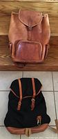 Two backpacks side by side; one black fabric with leather straps and buckles and the other brown leather knapsack, both visible from front