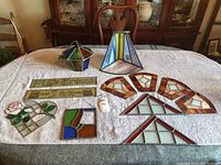 Photo of the stained glass pieces spread on a white cloth, showing various geometrical and floral stained glass panels plus a small hanging multicolored piece in purple, green, and white
