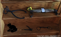 Photo of five black metal tools and a wooden yoke arranged on wooden steps; tools include large ice tongs, horse bit, smaller tongs, and a hand-forged hook tool.