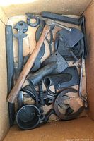 View of box containing assorted vintage metal tools and hardware including stone pick with wooden handle, wrenches, horseshoe, and metal castings.