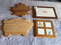 Four pieces displayed including a maple leaf shaped wooden board, pig shaped wooden board, wooden tray with a floral print, and wood framed ceramic tile trivet with botanical tiles.
