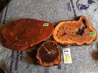 Three live edge wood wall clocks placed on the floor, showing natural wooden textures and edges. One clock is missing hands. The clocks vary in size and wood grain color.