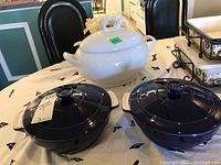 Photo showing large white ceramic casserole with lid and ladle, alongside two navy blue round casserole dishes with lids on a table.