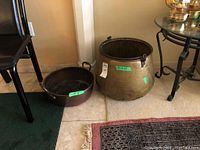 Two large vintage copper pots side-by-side on tiled floor showing relative sizes and conditions, including dents and wear on the exterior surfaces.