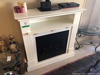 Front and side view of white electric fireplace with mantle shelf, black fireplace insert with glass doors, placed against a wall with decor items on top and nearby.