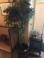 Full view of the faux plant with dense green leaves in large dark ceramic pot beside furniture.
