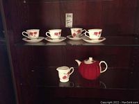 Red ceramic teapot covered with a red knitted cozy with matching ceramic creamer with red rose floral pattern, displayed on dark glass shelf.