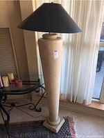 Full view of the pedestal floor lamp showing beige tapered base and black conical shade, positioned next to a glass and metal table and window.