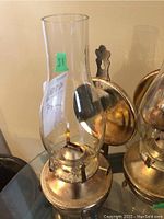 Two brass kerosene lamps with clear glass chimneys and round mounting plates placed on a reflective surface.