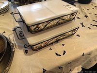 Two Temptations ceramic serving dishes stacked, showing floral patterned bases and white lids with wire racks.