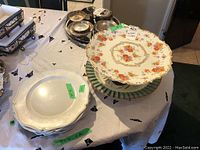 Porcelaine de France hand-painted cake holder with floral motif on top of Ceramica Due Torri pasta bowl and several silver plates and chargers on a table with a white cloth.