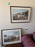 Overall view of two framed prints leaning against wall above couch, showing both battle scenes in worn but intact frames.