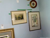Two framed art prints hanging on a wall. One depicts a floral arrangement and the other a village scene. Both have gold-tone frames and are signed by the artist.