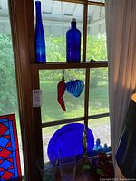 Blue glass bottles, a blue glass heart hanging ornament, blue plate excluded, and a blue glass decanter on a window sill with outdoor background.