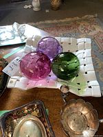 White woven ceramic bowl holding three blown glass balls in pink, purple and green. Surrounding silver and other decorative items visible.