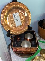 Hammered brass plate displayed on a stand with copper bowl, two mugs, and rectangular container below it.