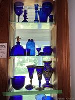Three shelves of assorted cobalt blue glassware photographed on wooden shelving near window, includes bottles, goblets, cups, vase, and candle holders