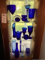 Full view of three shelves with various cobalt blue glass items arranged including goblets, vases, a paperweight, and candle holders.