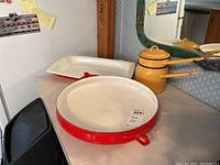 Overview of the Dansk red enamel round roasting dish, white rectangular roasting dish, and Polish yellow double boiler set on countertop with mirror background.