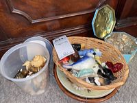 Wade and bird figurines in a basket and a plastic container on a wooden surface with some plates and trays nearby.