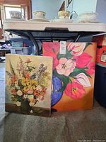 Two vintage floral paintings on canvas leaning against a table, one with a mixed bouquet in a dark vase and another of tulips in a blue vase.