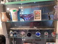 Shelf display showing a collection of seven small blown glass globes or paperweights each with unique colorful internal designs, alongside decorative glass vases and animal figurines.