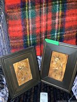Two framed wood carvings displayed side by side, showing detailed female figures and dark wood frames, against a plaid textile background.