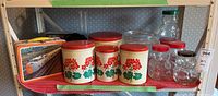Shelf with vintage tins with red lids and floral design, CN train lunch box, clear plastic bear-shaped jars, napkin holder, and a large glass bowl.