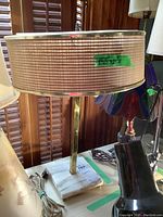 Mid Century Modern style table lamp on a shelf, featuring a white square marble base, vertical metal rod, and round beige woven fabric lampshade with gold trim.