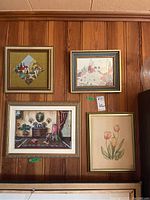 Photo of four framed needlepoint artworks hung on wood panel wall showing floral, marine, and interior scene themes.