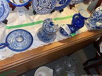 Blue and white ceramic items including lidded jar, candlestick holder, pitcher, and bowl arranged on a wooden surface with a lace table runner.