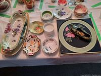 Overview of assorted decorative ceramic and porcelain plates, small dishes, a ceramic shoe, and a cup with saucer on a table.