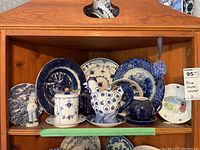 Full shelf showing eight blue and white plates, a teapot, sugar bowl, ceramic chicken figurine, cup, and bowl arranged on wood shelf.