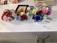 Three porcelain flower arrangements grouped on a white surface showing details and condition.