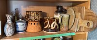 Shelf showing various pottery items including a mottled brown and black bowl, orange terracotta holder, green-tinted pottery pitcher, and wooden cutting boards