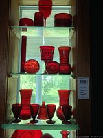 Three shelves of ruby red glassware displayed on glass shelves in a window, including various vases, goblets, bowls and decorative spheres.