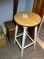 Round wood stool with natural finish seat and white legs in front of small oak shoe shine box bench showing wear.